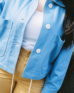 A person wearing a light blue denim jacket over a white top and tan drawstring pants, shown from the chest down in a close-up lifestyle shot.
