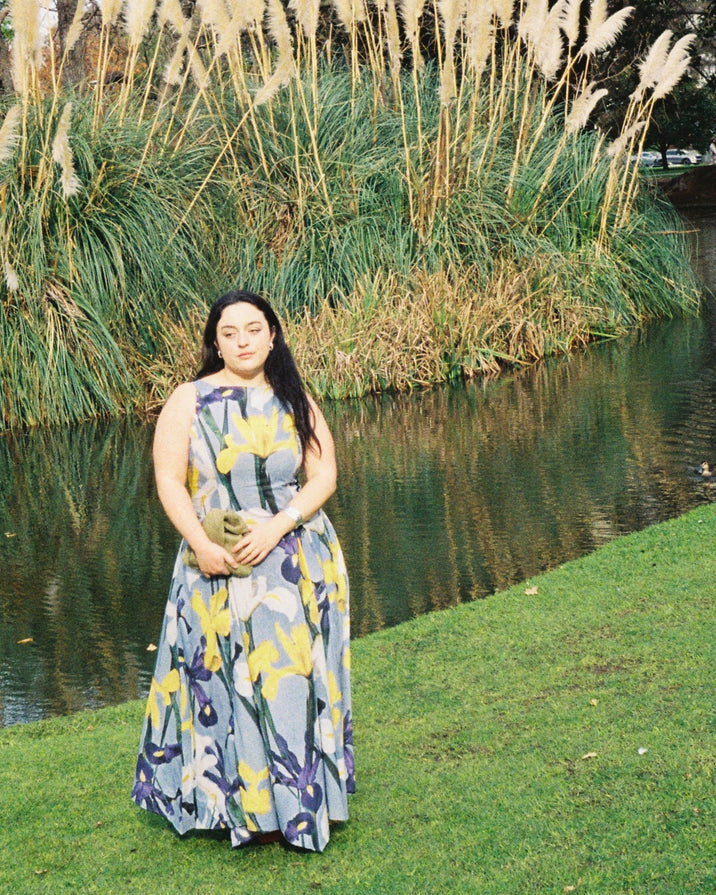 A woman stands outdoors by a pond with tall reeds and grass in the background, wearing a sleeveless maxi dress with a blue-gray base and a yellow and purple abstract floral pattern. She is holding a small olive-green clutch.