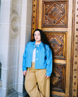 A woman stands in front of a large ornate wooden door wearing a bright blue cropped denim jacket over a white top and tan wide-leg pants.