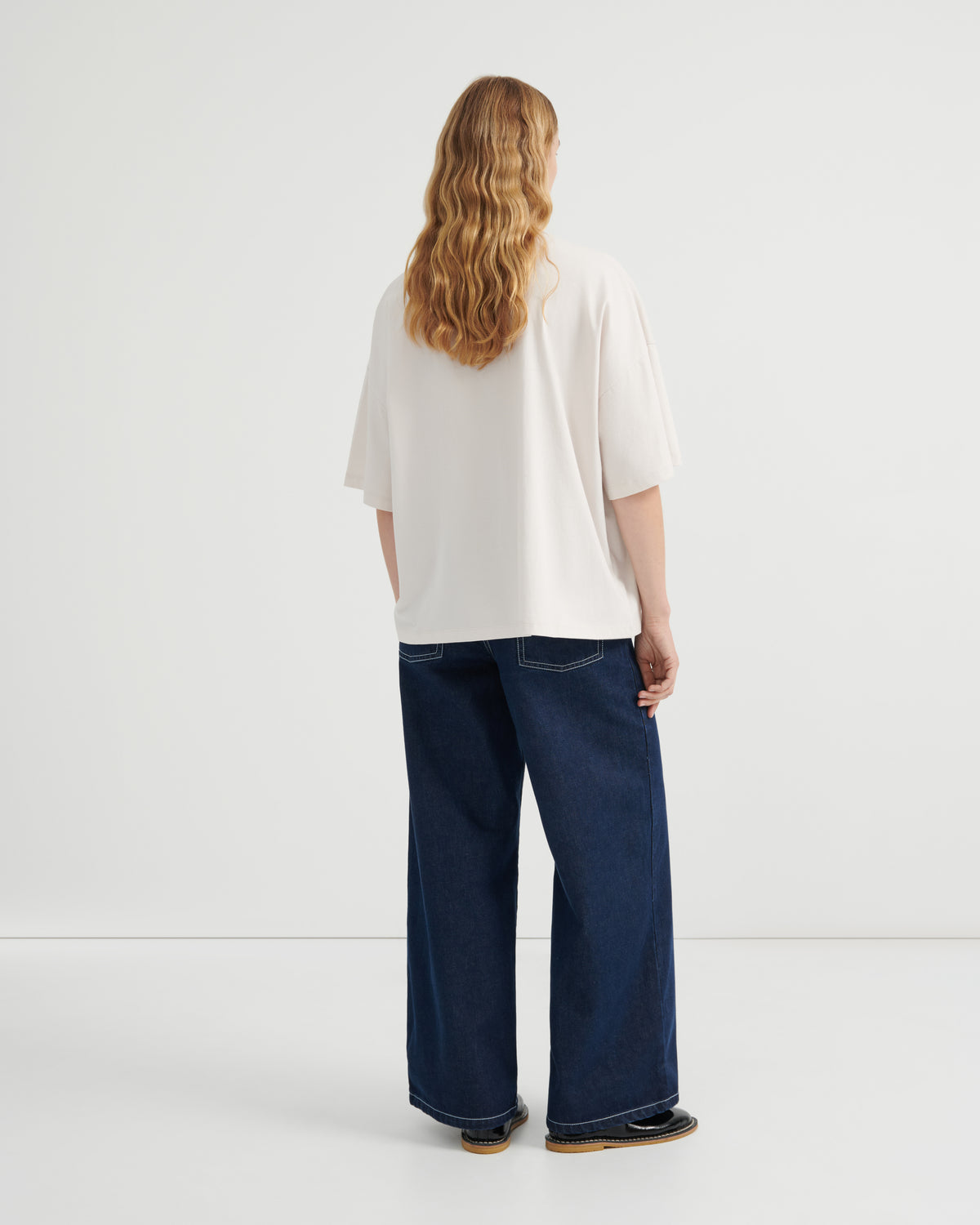 A woman with long wavy light brown hair is shown from behind wearing an oversized off-white short-sleeve top and dark blue wide-leg jeans, standing against a clean white studio background.