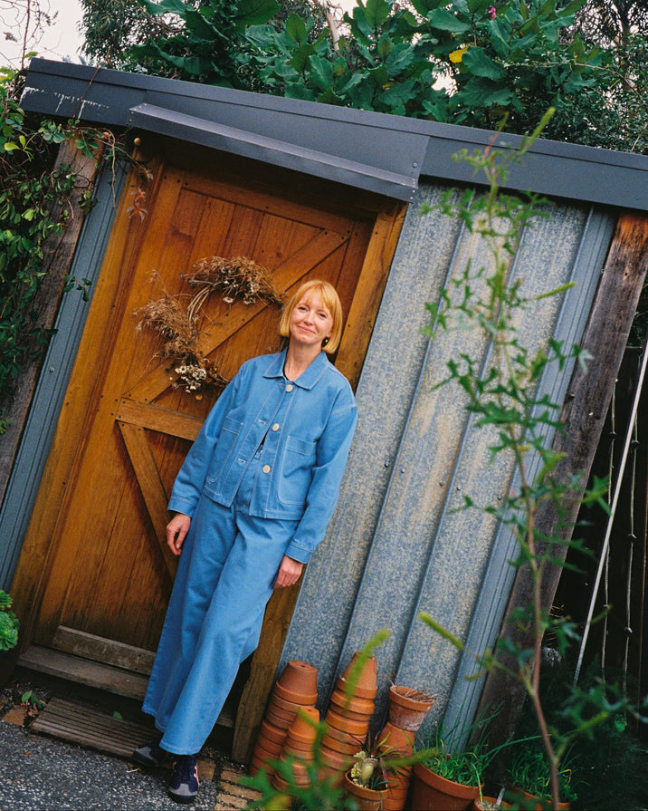 A woman with short blonde hair wearing a blue denim jumpsuit stands in front of a rustic wooden door of a corrugated metal shed, surrounded by green garden foliage and potted plants.
