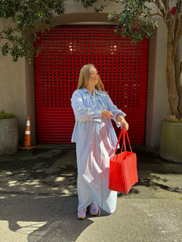 A woman seen from behind wearing a light blue and white vertically striped shirt dress, carrying a large red woven tote bag against a dark brown wall.