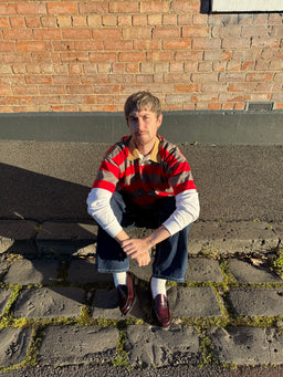 A man crouching outdoors on a stone pavement in front of a brick wall, wearing a red, brown, and beige striped polo shirt over a white long-sleeve, dark blue jeans, white socks, and burgundy loafers.