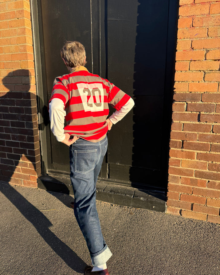 A person standing outdoors in front of a brick building and dark door, wearing a red and beige striped long-sleeve sweater with a large white number 20 on the back, blue jeans, and brown shoes.