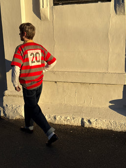 A person walking outdoors in warm sunlight wearing a red and gray striped long-sleeve jersey with the number 20 on the back, over a white long-sleeve shirt and dark jeans.