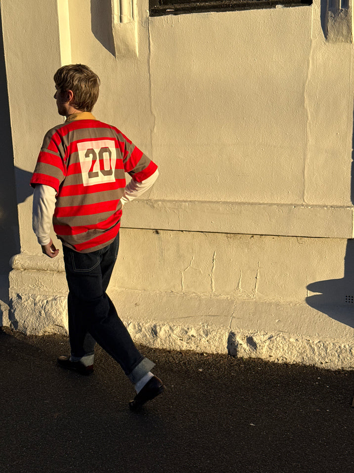 A person walking outdoors in warm sunlight wearing a red and gray striped long-sleeve jersey with the number 20 on the back, over a white long-sleeve shirt and dark jeans.
