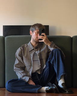 A man takes a mirror selfie while sitting on a dark green couch. He is wearing a brown and beige vertically striped button-down shirt over a white undershirt, dark blue jeans, white socks, and black leather loafers.