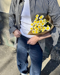 A person outdoors wearing a black-and-white vertically striped button-up shirt over a white t-shirt, blue jeans, and black shoes, while holding a bouquet of yellow and white daisies wrapped in brown paper.