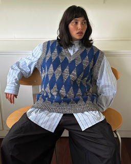A model sits on a wooden chair wearing a blue and gray argyle knit sweater vest over a light blue and white striped button-down shirt, paired with wide-leg black trousers.