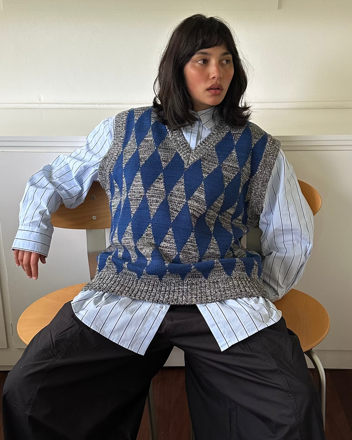 A model sits on a wooden chair wearing a blue and gray argyle knit sweater vest over a light blue and white striped button-down shirt, paired with wide-leg black trousers.