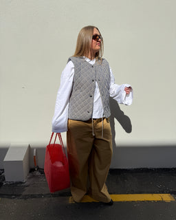 A woman stands outdoors against a plain light wall wearing a gray and white patterned vest over a white long-sleeve blouse, tan wide-leg pants, and black sunglasses. She is holding a bright red tote bag.