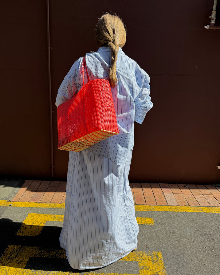 A woman seen from behind wearing a light blue and white vertically striped shirt dress, carrying a large red woven tote bag against a dark brown wall.