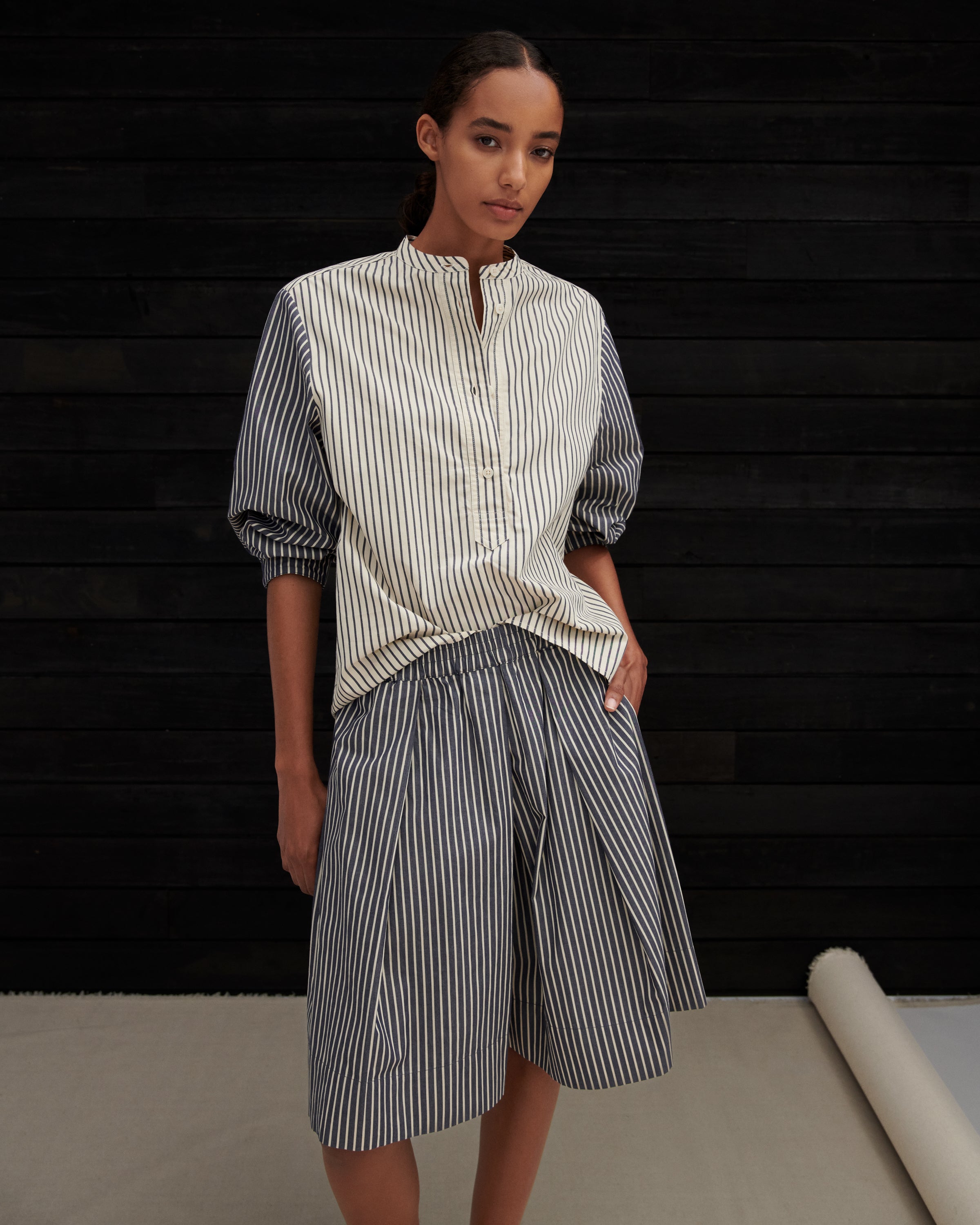 A model wearing a striped shirt and matching striped shorts, standing against a dark wood-paneled wall.