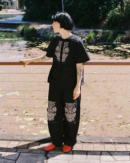 A person standing outdoors near a pond wearing a black short-sleeve top and matching loose pants with ornate white embroidered or printed patterns on the chest and upper legs, paired with bright red shoes.