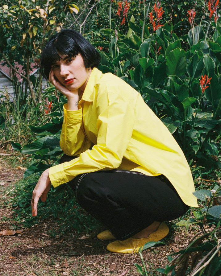 A person crouching outdoors in a garden, wearing a bright yellow button-up shirt, black pants, and yellow shoes.