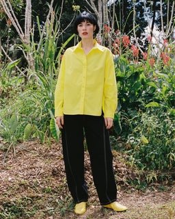 A model stands outdoors in a lush garden setting, wearing a bright yellow long-sleeve button-up shirt, black wide-leg trousers with white piping, and matching yellow shoes.