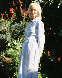 An older woman with white hair stands outdoors in a garden wearing a long-sleeved, belted maxi dress with thin vertical light blue and white stripes. The background consists of lush green foliage and small red flowers.