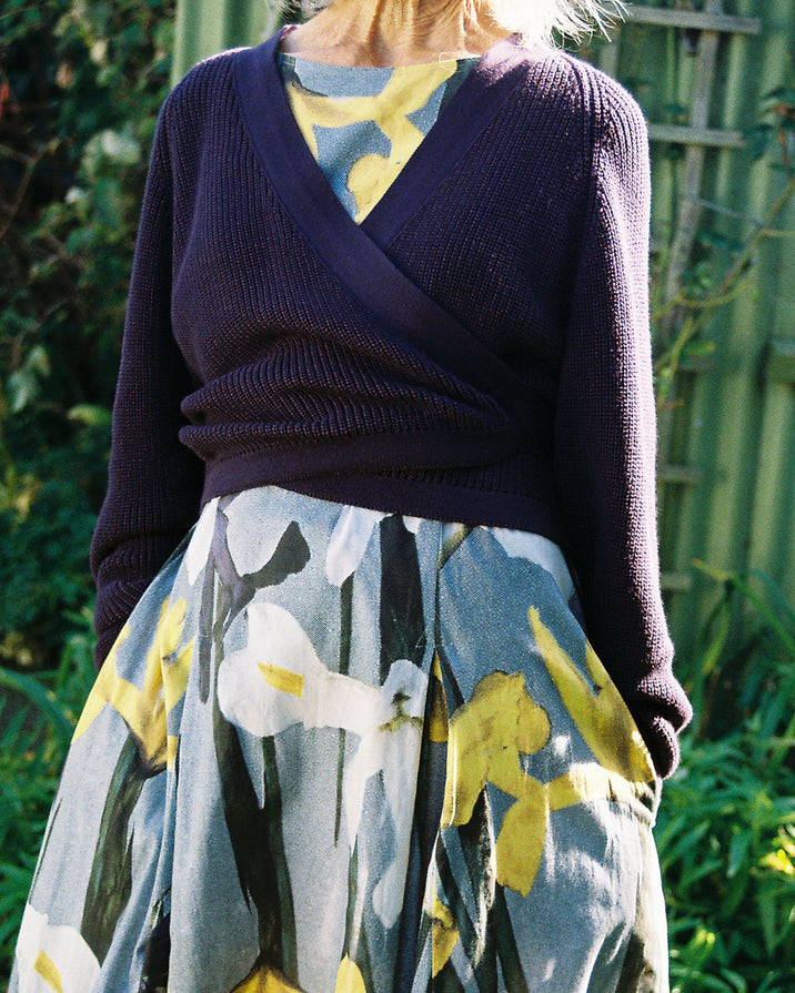 A person outdoors wearing a dark purple wrap-style knit sweater over a yellow and blue patterned top, paired with a gray skirt featuring yellow and white floral motifs.