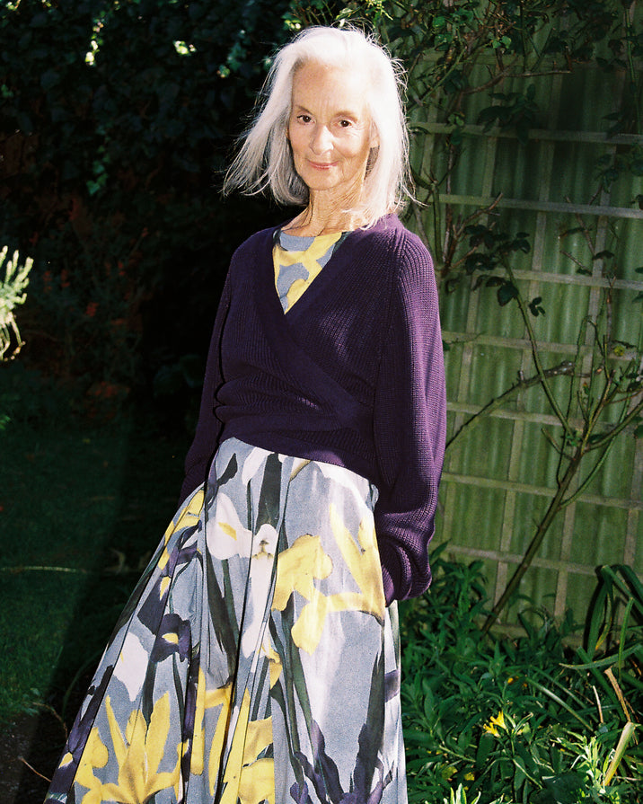 An elderly woman with white hair stands outdoors in a garden wearing a purple knit wrap sweater and a long pleated skirt with a yellow, gray, and green floral pattern.