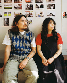 Two people sitting side by side indoors, photographed against a wall covered with small photos. The person on the left wears a white T-shirt, blue and gray argyle sweater vest, and light khaki pants.