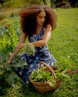 A woman with voluminous curly hair sits in a grassy garden, harvesting green broccoli from a plant while wearing a blue and white patterned dress. A wicker basket filled with leafy greens sits beside her.