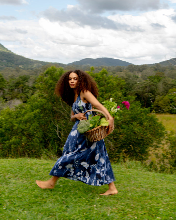 A woman with long curly hair walks barefoot across a grassy field, wearing a blue and white tie-dye maxi dress and carrying a wicker basket filled with green vegetables and pink flowers, with mountains and a cloudy sky in the background.