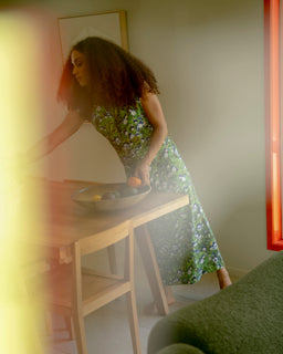 A woman with curly hair wearing a green floral print midi dress leans over a light wood dining table, placing fruit into a bowl. The photo is shot from an off-center perspective with a blurry green foreground element and warm, hazy lighting.