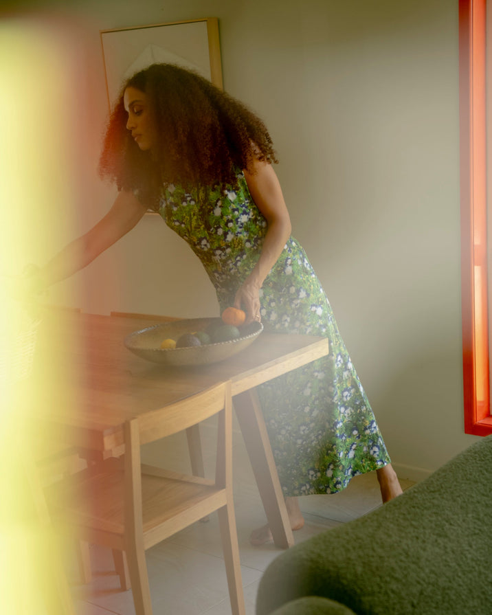 A woman with curly hair wearing a green floral print midi dress leans over a light wood dining table, placing fruit into a bowl. The photo is shot from an off-center perspective with a blurry green foreground element and warm, hazy lighting.