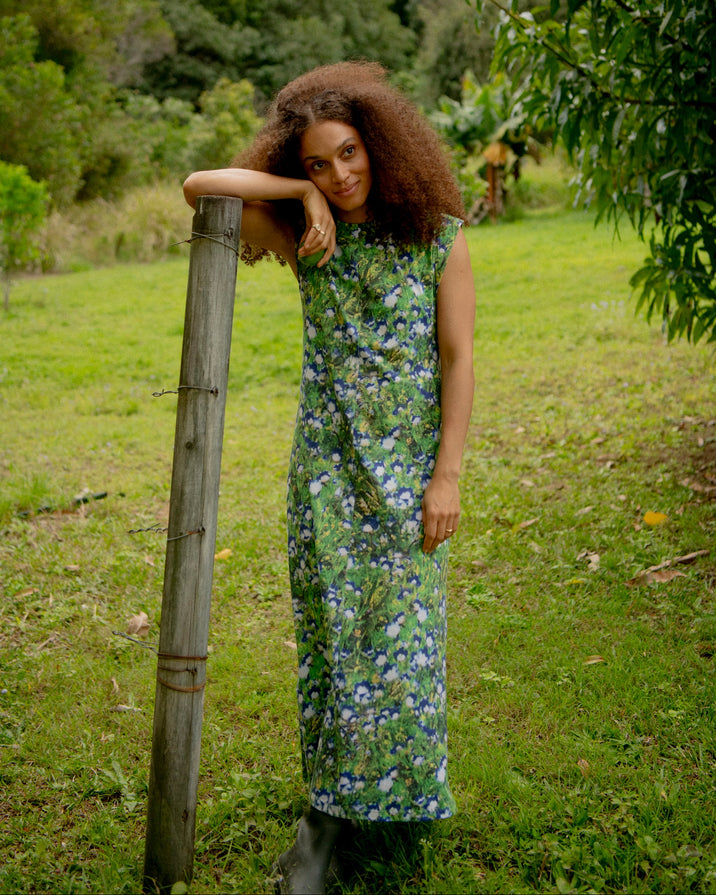 A woman with curly brown hair stands outdoors in a grassy field leaning against a wooden post, wearing a sleeveless, knee-length green dress with a blue and purple floral pattern.
