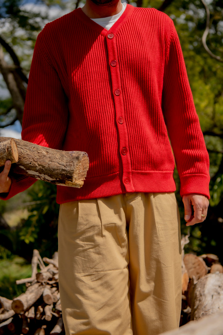 A person outdoors wearing a bright red ribbed button-front cardigan over a white undershirt and beige pants, holding a log. The background shows trees and stacked firewood in natural daylight.