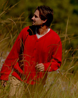 A man wearing a bright red knit cardigan over a white undershirt and tan pants, standing in tall sunlit grass outdoors.