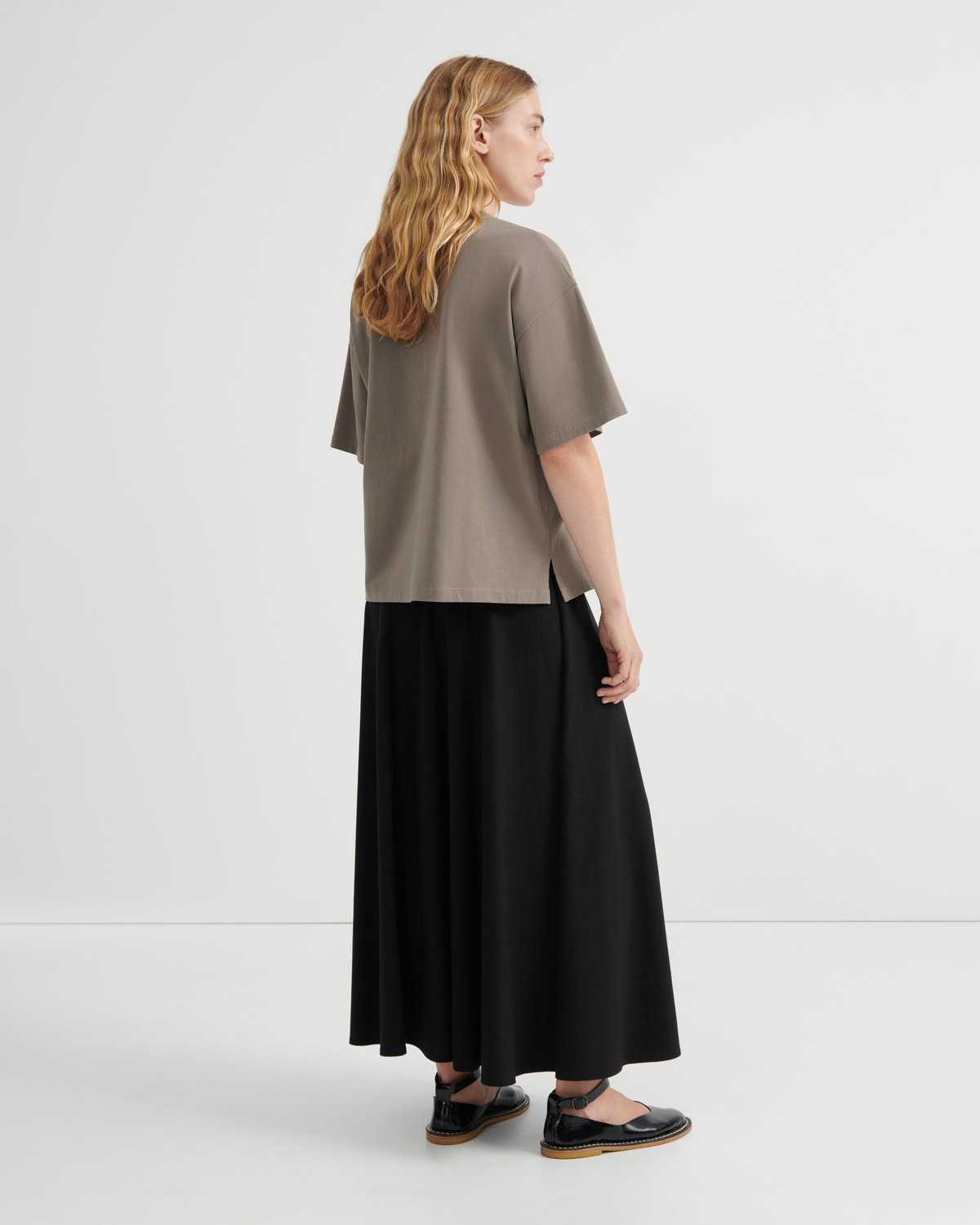A woman stands in profile wearing an oversized taupe short-sleeve top and a long black skirt, photographed in a clean studio setting.