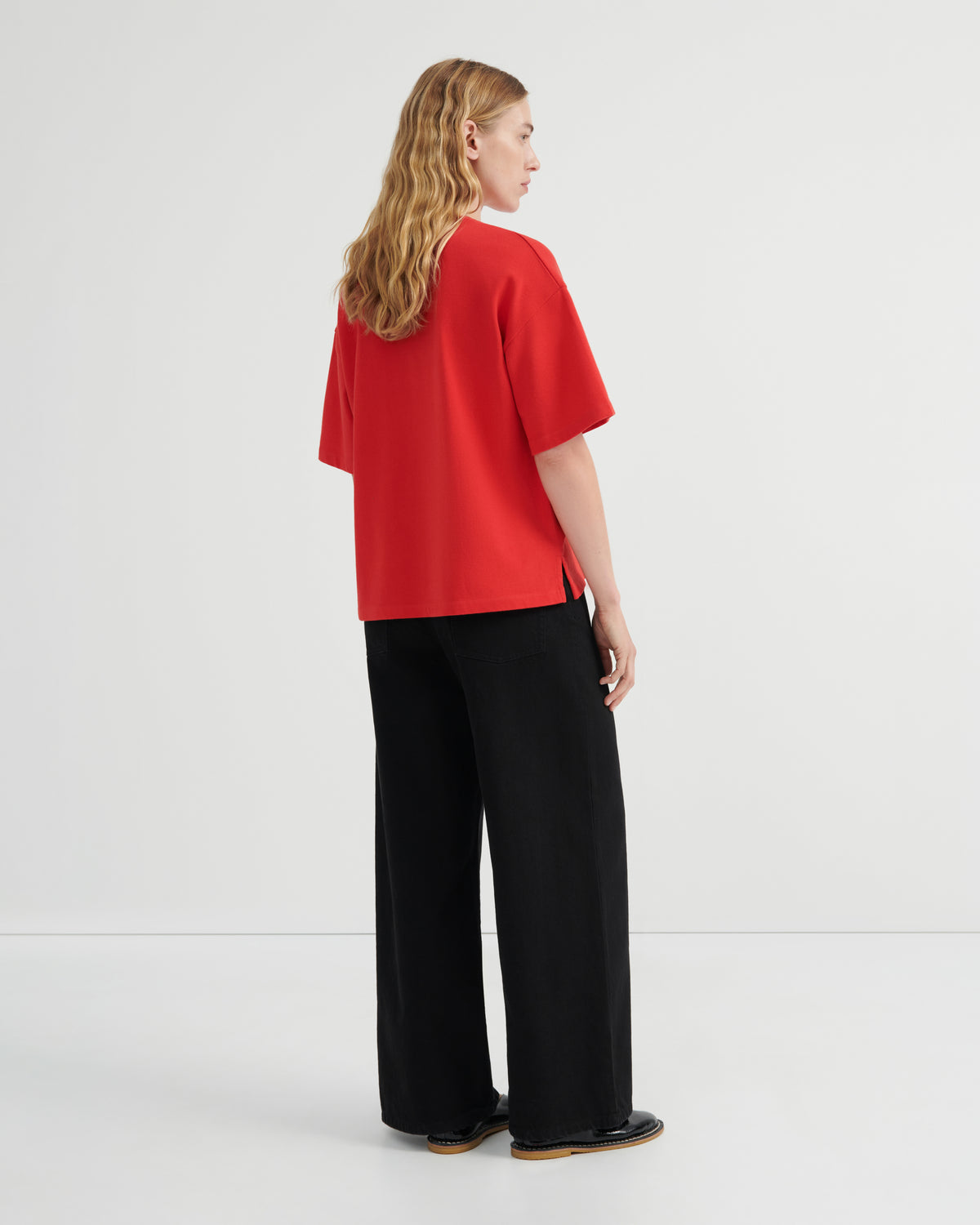 A woman stands in profile wearing a bright red oversized short-sleeve top and black wide-leg trousers against a clean white studio background.