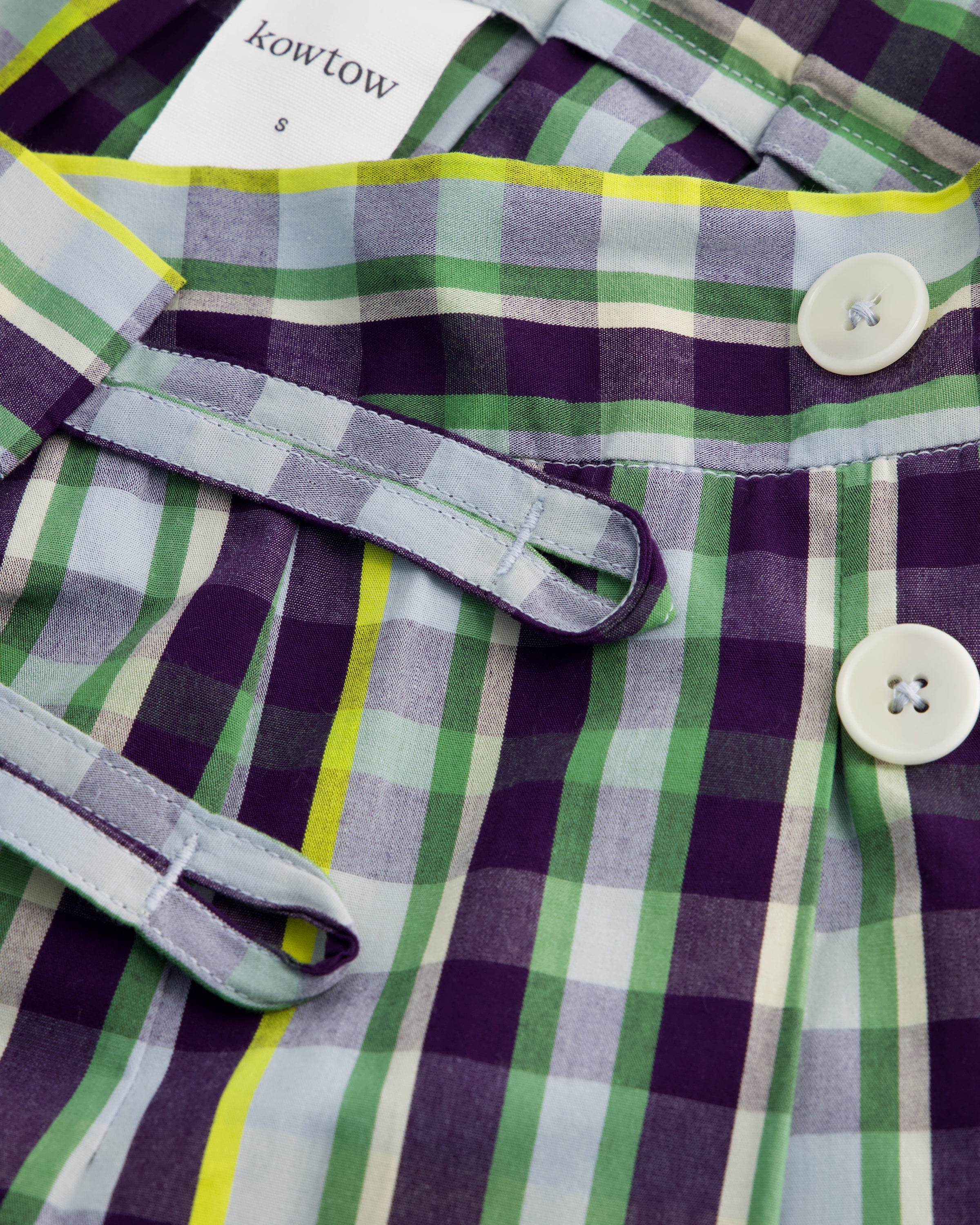 Close-up of a Kowtow plaid garment, likely a skirt or dress, featuring a purple, green, and white check pattern with a lime green stripe. The image shows the waistband with white buttons and a matching fabric tie belt.