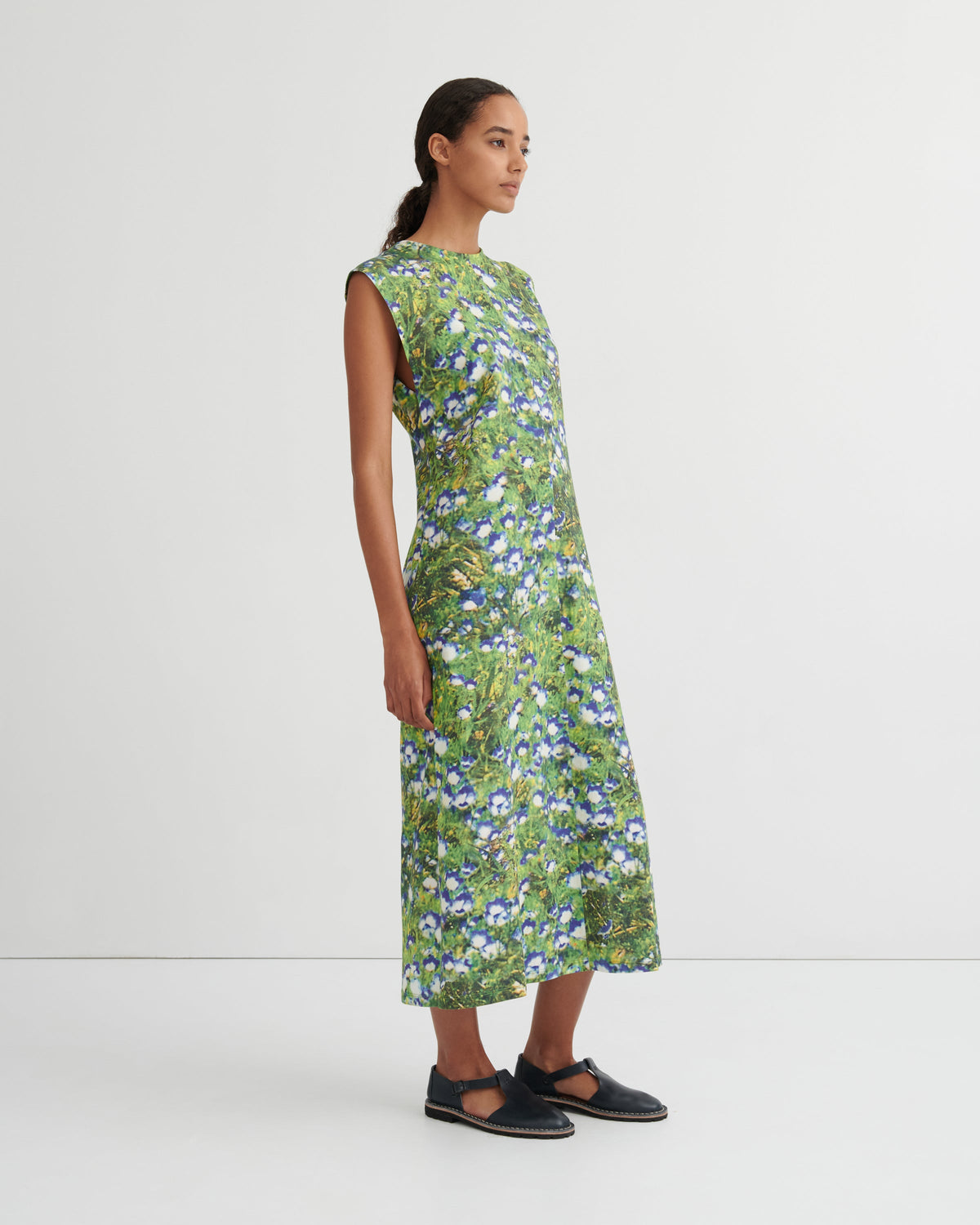A woman modeling a sleeveless, knee-length green dress with a dense blue and white floral print, paired with black Mary Jane flats, against a plain white studio background.