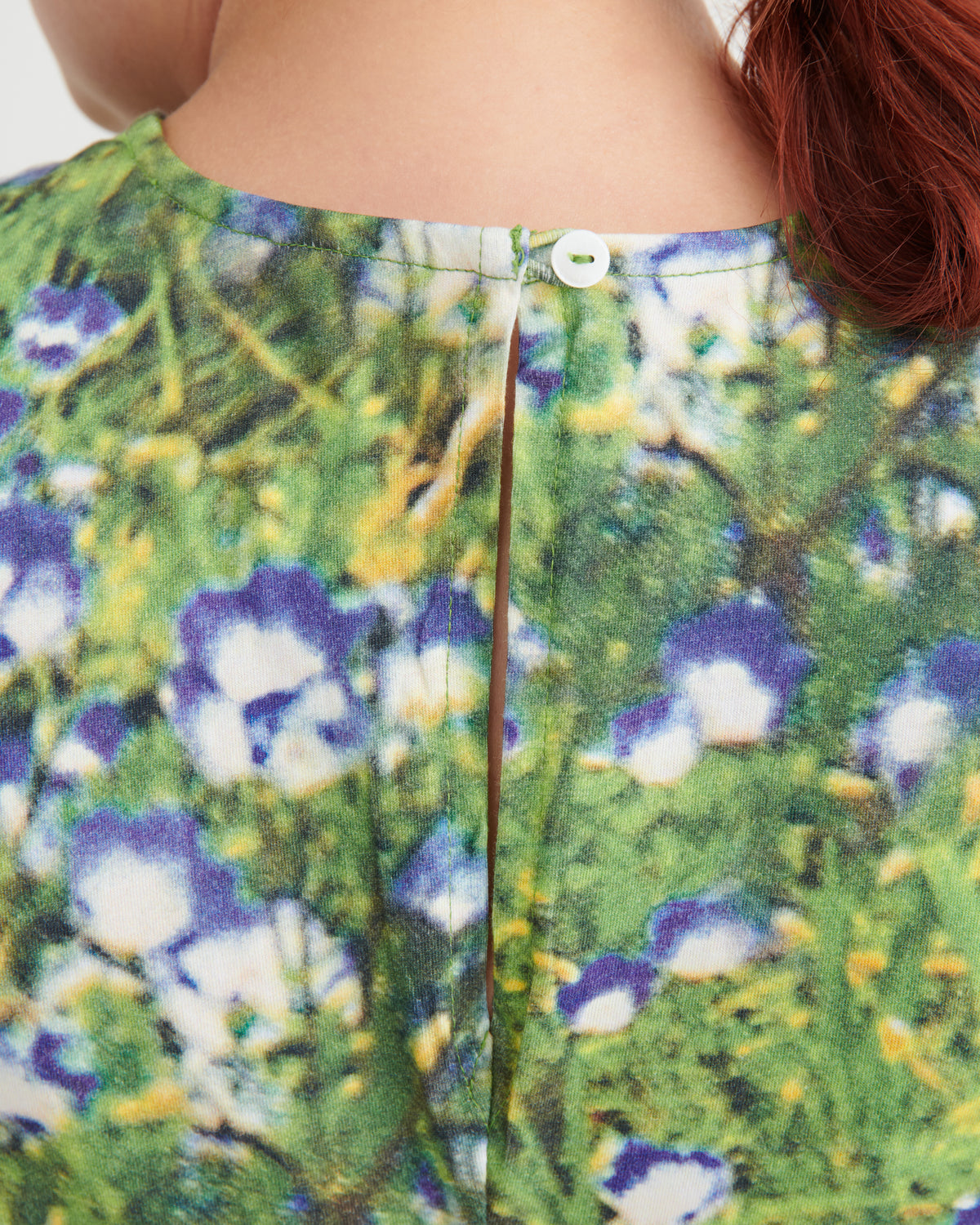 Close-up back view of a person wearing a floral print top with green foliage, purple flowers, and white blossoms, featuring a central seam and a small white button at the neckline.