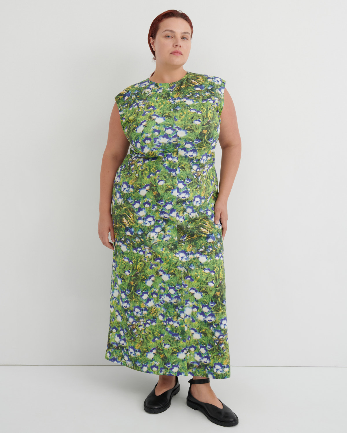 A woman modeling a sleeveless, knee-length maxi dress with a green and blue floral print, paired with black flat shoes, against a plain light gray studio background.