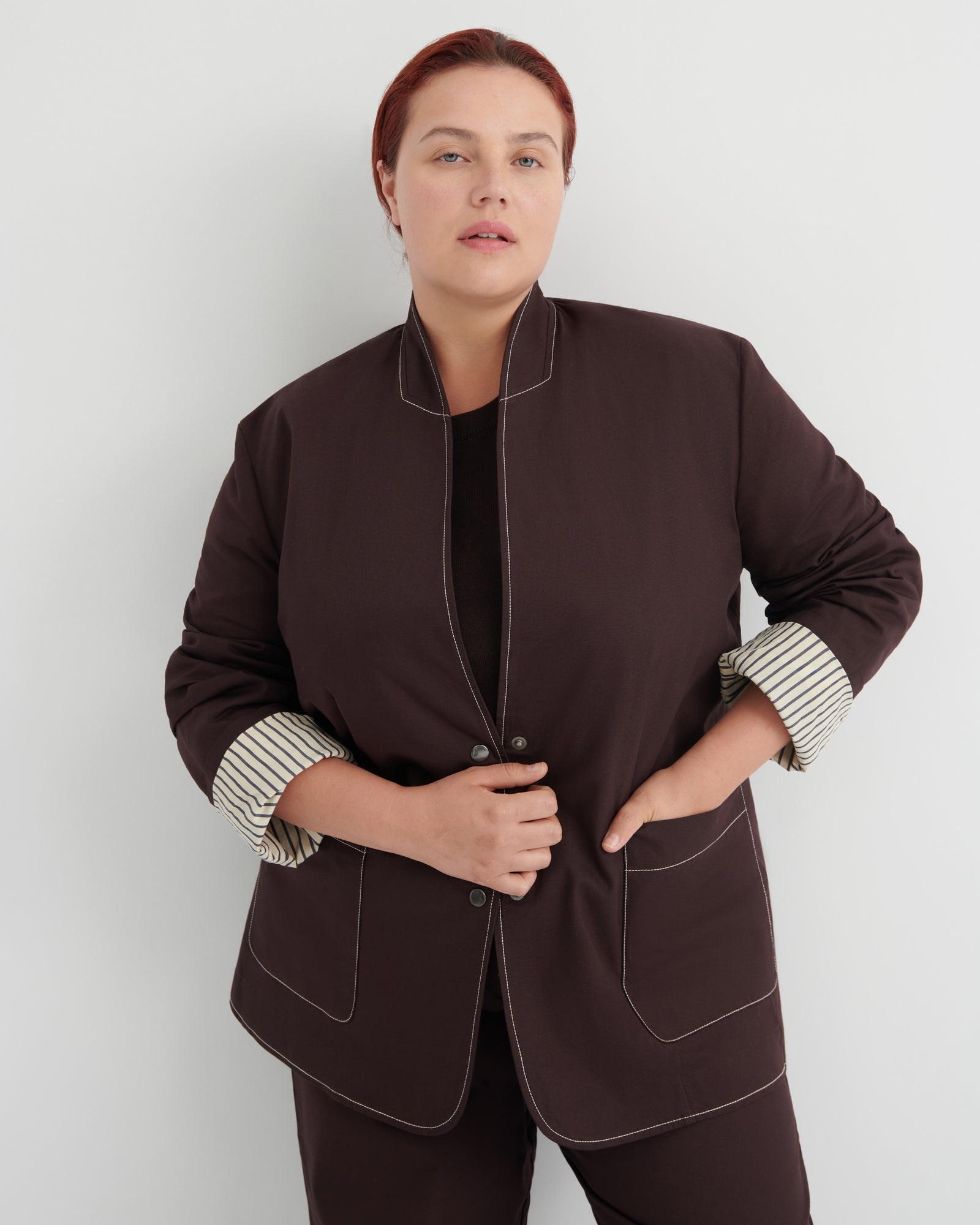 A woman modeling a dark brown jacket with contrasting white topstitching and striped button-up cuffs, paired with matching dark brown trousers against a light gray studio background.