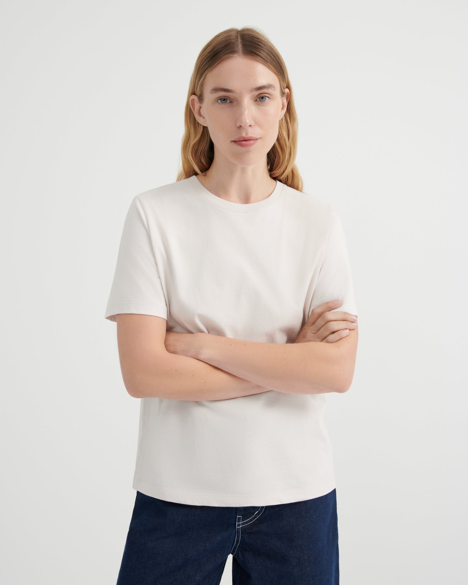A woman with blonde hair wearing a plain off-white short-sleeve crewneck t-shirt and dark blue jeans, standing against a light gray studio background with her arms crossed.