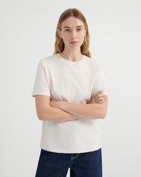 A woman with blonde hair wearing a plain off-white short-sleeve crewneck t-shirt and dark blue jeans, standing against a light gray studio background with her arms crossed.