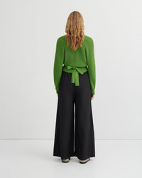 A woman viewed from the back wearing a bright green long-sleeve knit top with a tied waist and black wide-leg trousers, standing against a plain light gray studio background.
