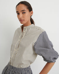 A woman wearing a cream-colored blouse with thin dark vertical stripes and contrasting dark blue puff sleeves, photographed against a neutral light gray background.