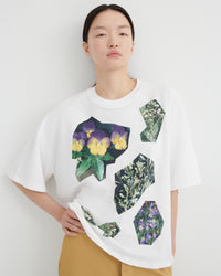 A model wearing a white oversized t-shirt with a collage print of purple and yellow pansies and green botanical patterns, paired with beige pants, against a plain light gray background.