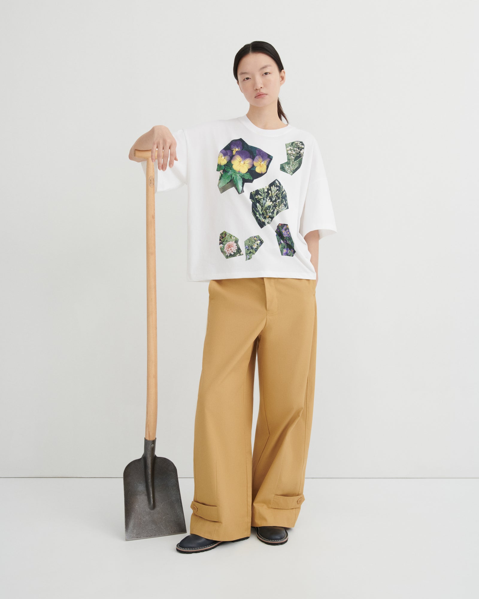 A model stands in a studio wearing a white graphic T-shirt with colorful abstract floral prints, tan wide-leg pants, and black shoes, while holding a shovel against a clean off-white background.