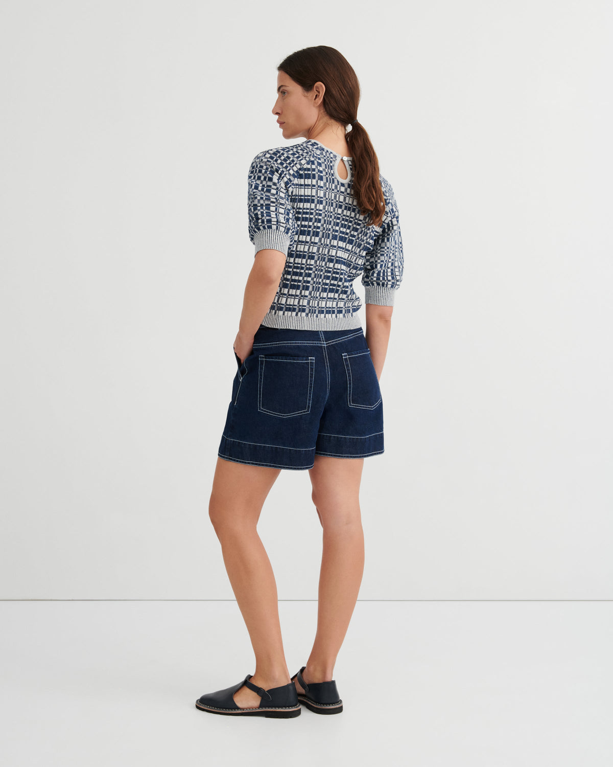 A woman is shown from the back wearing a short-sleeve knit top with a navy and white geometric pattern, dark blue denim shorts, and black sandals, set against a plain white studio background.