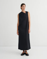A model wearing a sleeveless, black draped midi dress with a high neckline, paired with black flat shoes, standing against a plain white studio background.