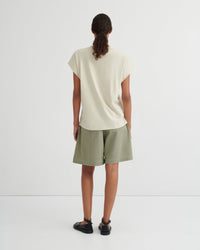 A model viewed from behind wearing a cream-colored short-sleeve t-shirt and sage green shorts against a plain white studio background.