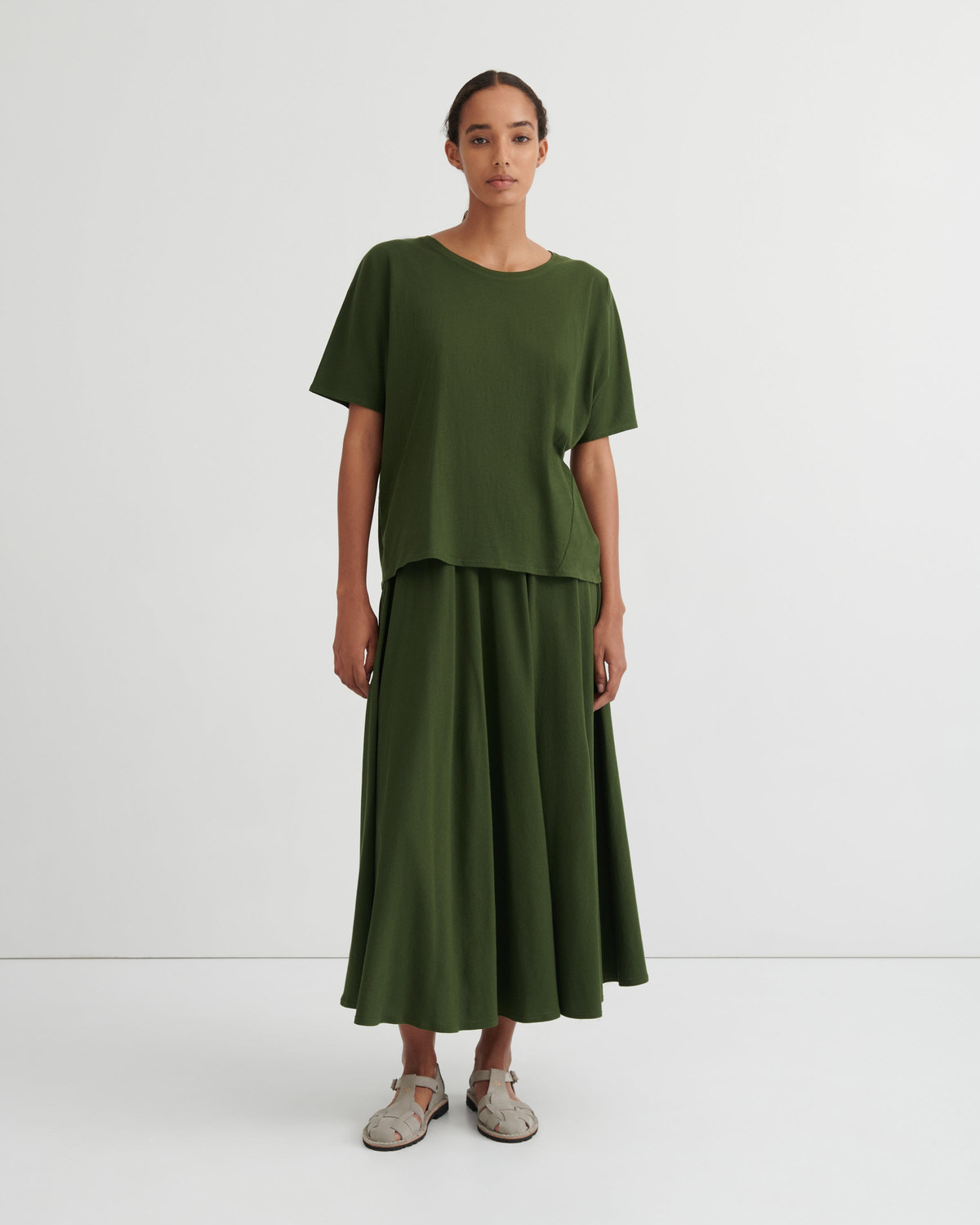 A model stands against a plain light gray studio background wearing an olive green two-piece outfit consisting of a short-sleeve crewneck top and a matching pleated maxi skirt, paired with light-colored flat shoes.