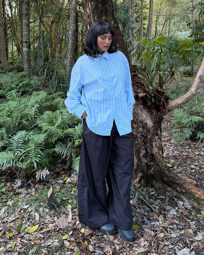 A woman stands outdoors in a wooded area wearing a light blue and white vertically striped button-up shirt and loose black wide-leg pants.