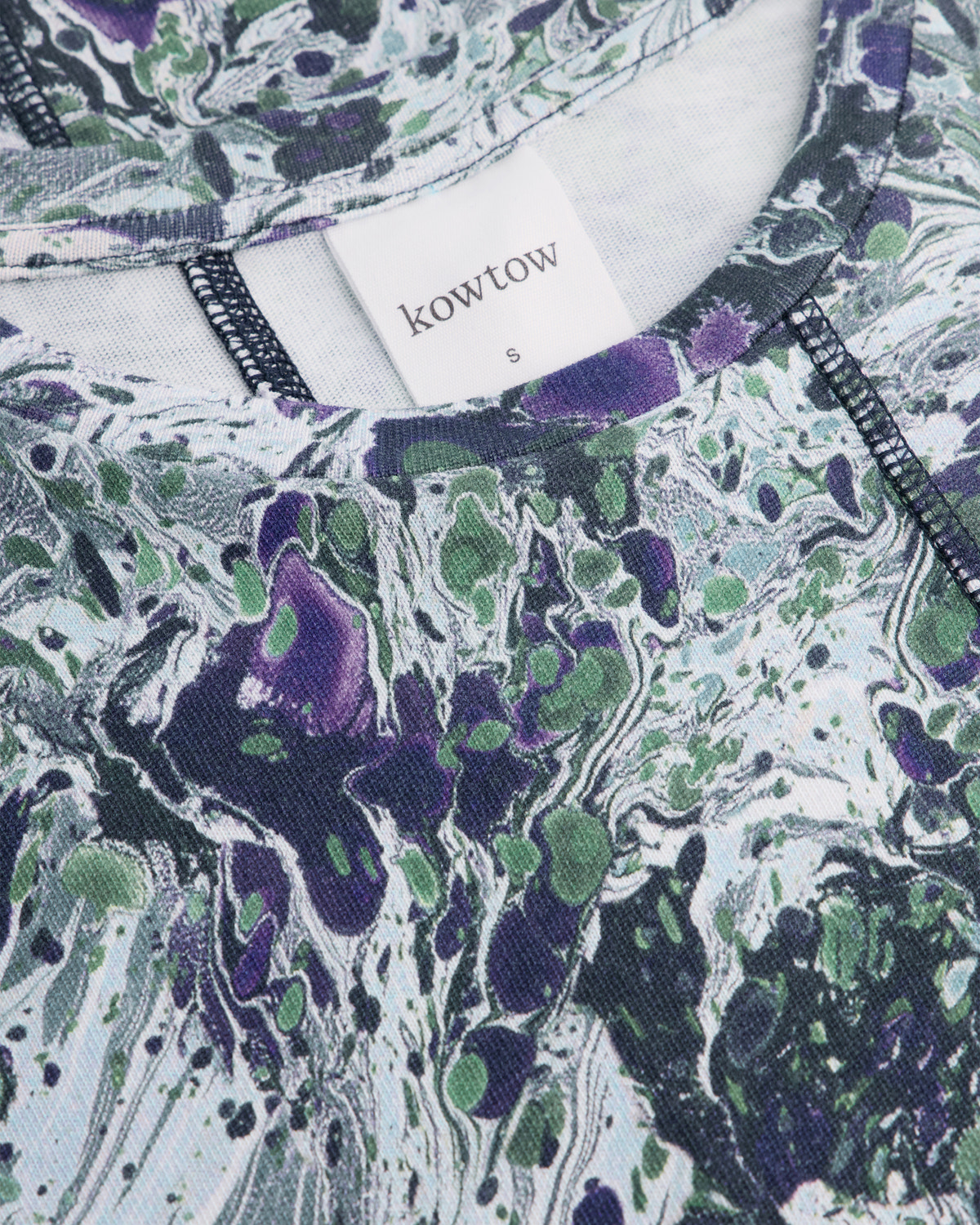 Close-up of a Kowtow garment featuring a marbled abstract pattern in purple, green, and white, with a white brand label visible at the neckline.
